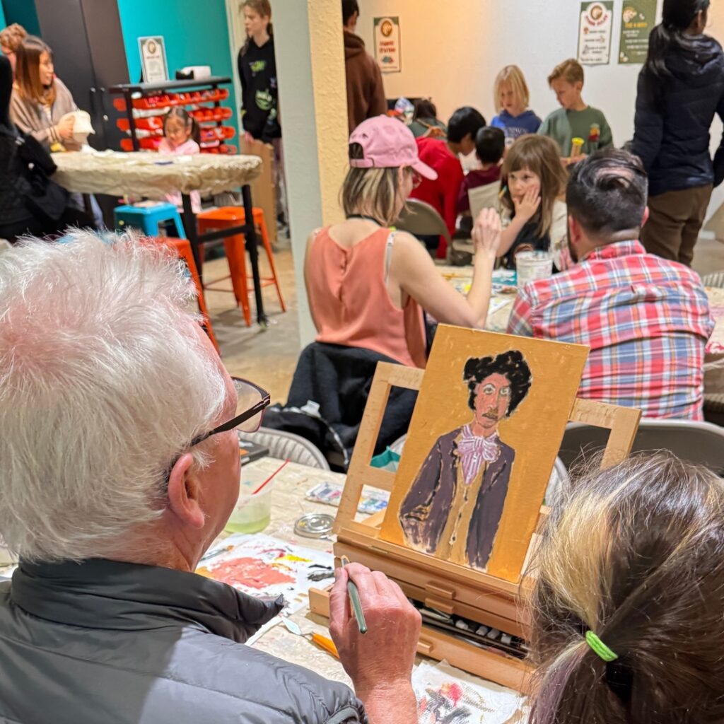 Participants of all ages paint together during a family open studio art session, with an instructor guiding a portrait painting in the foreground.