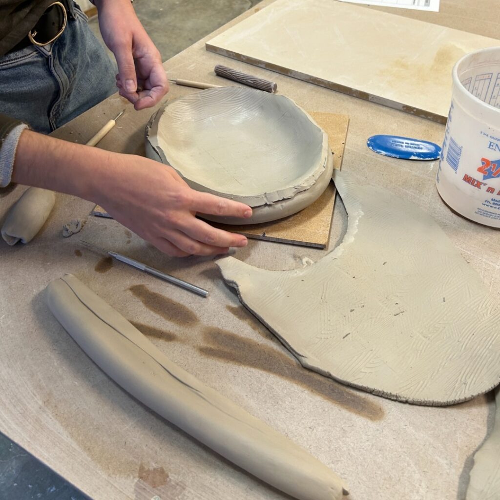 Hands shape and assemble a clay bowl using handbuilding techniques on a worktable with ceramic tools.