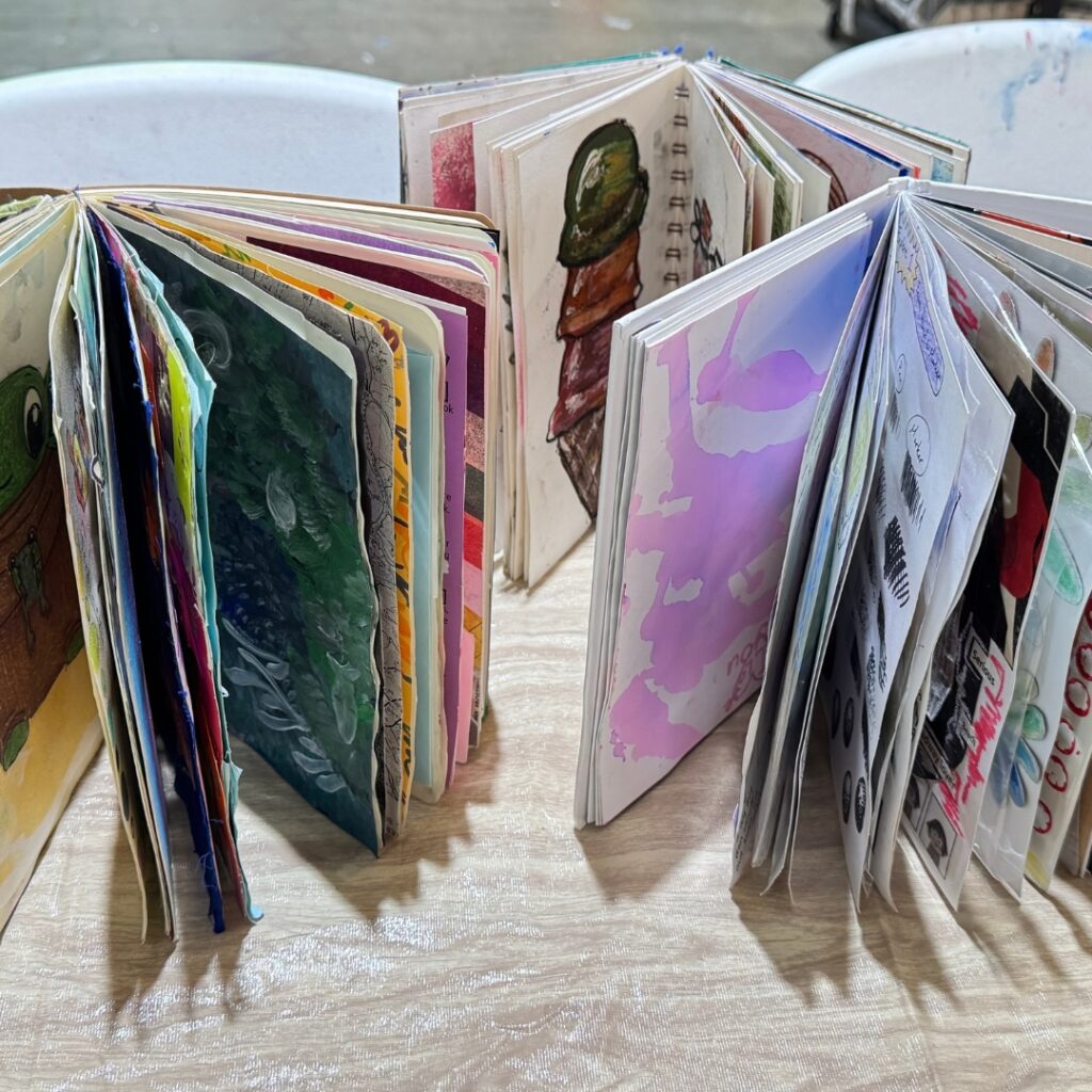 Handmade sketchbooks stand open on a table, displaying colorful mixed-media artwork and painted pages.