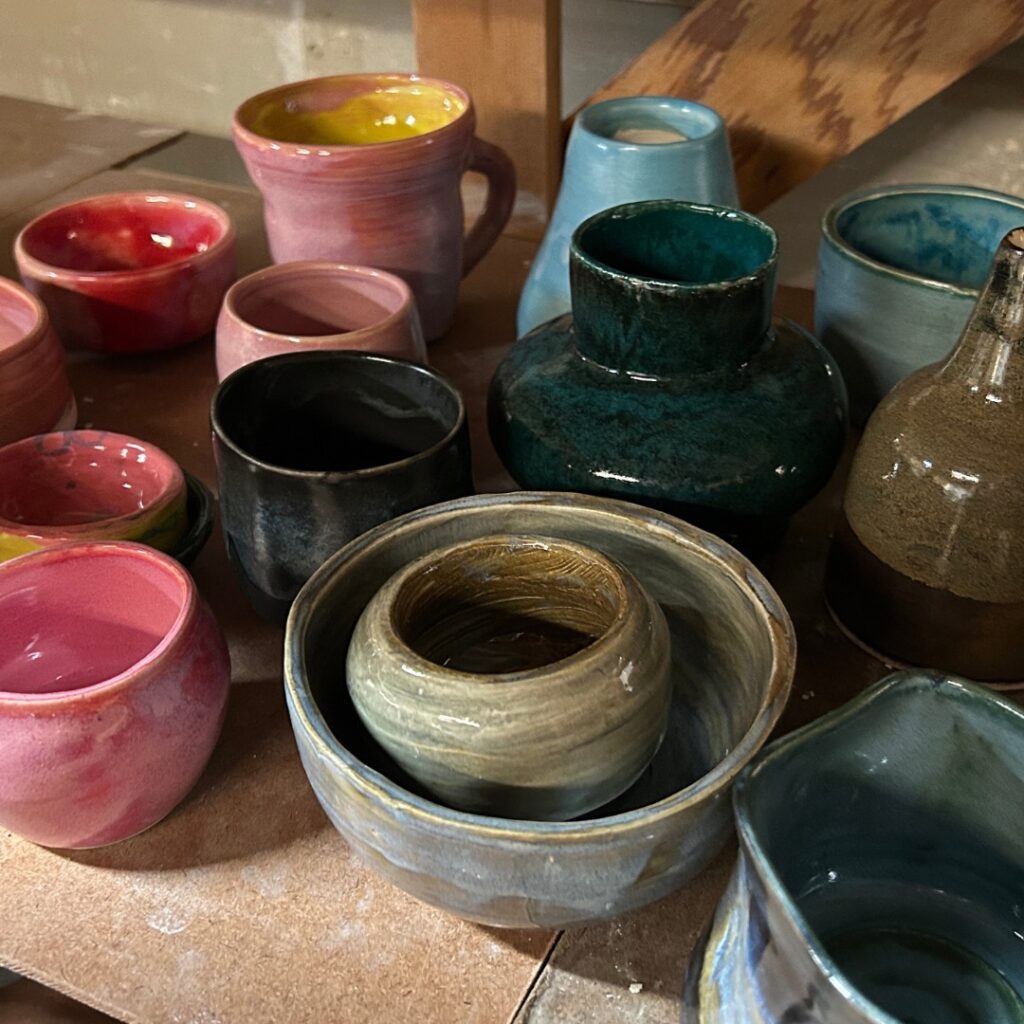 Colorful glazed ceramic bowls, mugs, and vessels made on the pottery wheel are arranged together on a studio shelf.