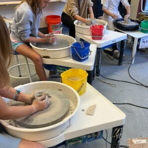 Children work at pottery wheels, shaping clay with their hands during a youth ceramics class.