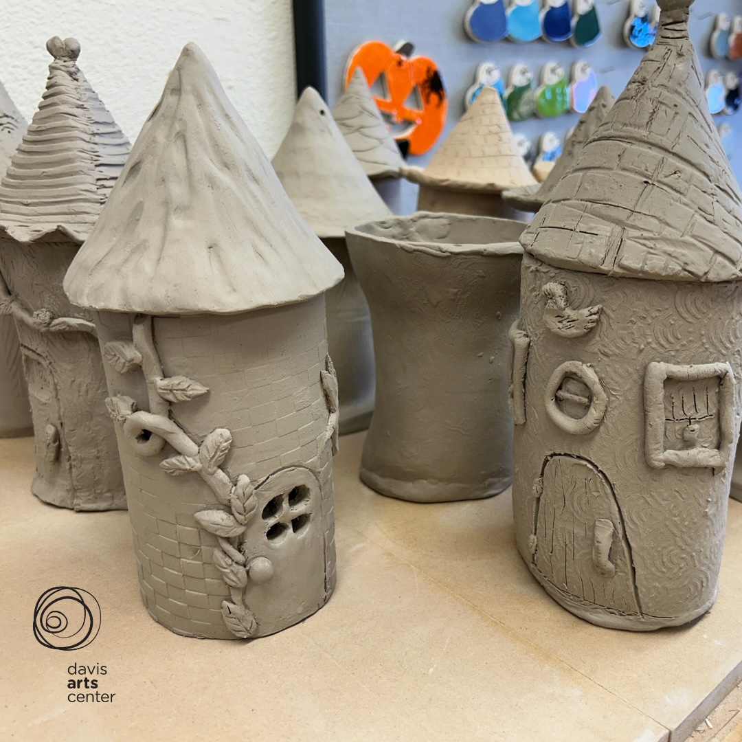 Small handbuilt clay houses with textured roofs, tiny doors, and leaf details displayed on a studio shelf.