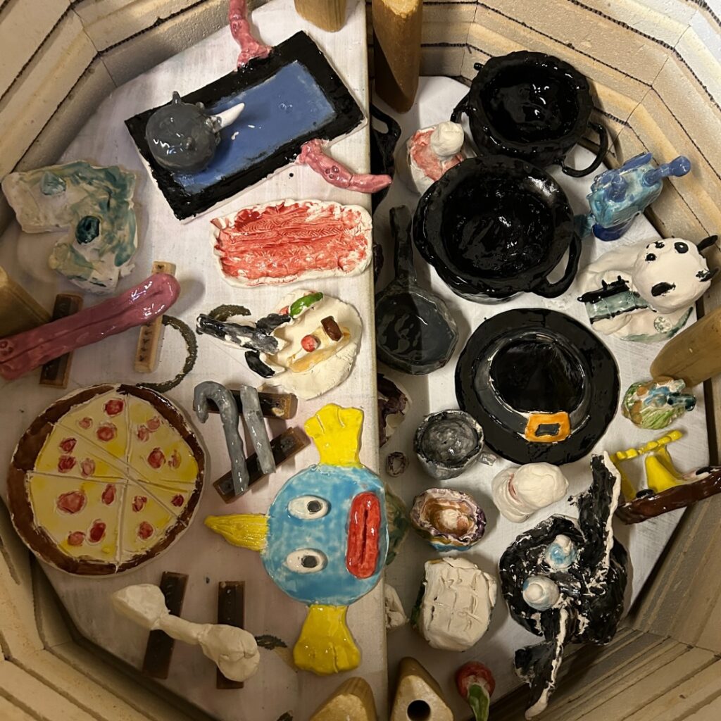 Assorted small glazed clay creations, including bowls, animals, food shapes, and whimsical figures, arranged inside a kiln.